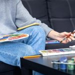 Person painting with watercolours at home, sitting on a sofa with art supplies on a table, wearing casual clothing